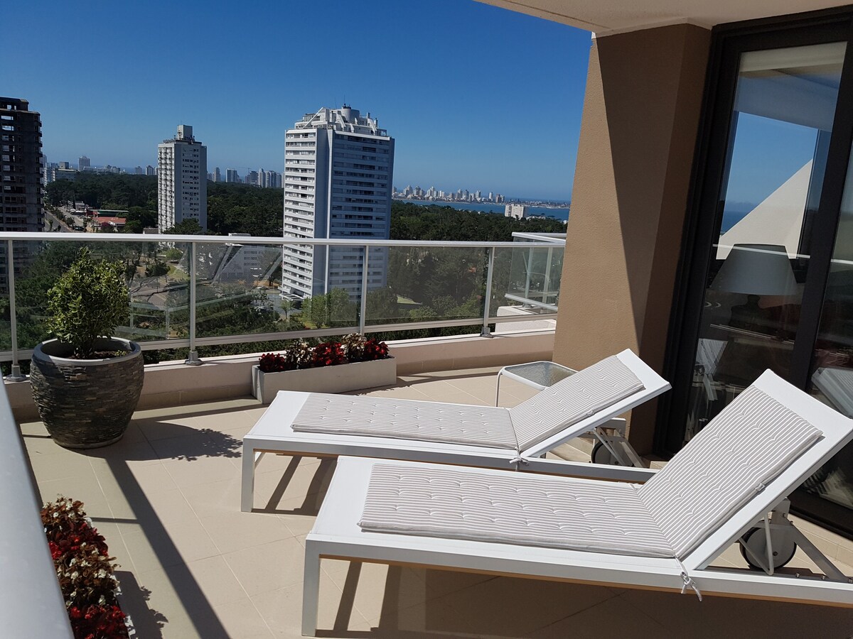 A spacious terrace features two white lounge chairs positioned for relaxation. Lush greenery and flower pots add color, while panoramic views of the surrounding buildings, forests, and the ocean can be observed in the distance under a clear blue sky.