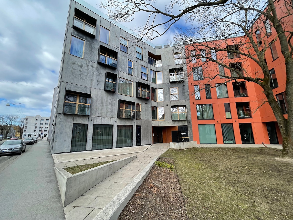 A modern apartment building exterior features a combination of grey and orange facades. Large windows are present, offering glimpses of the inside. A tree is visible along the side, and a small landscaped area with grass lies in front.