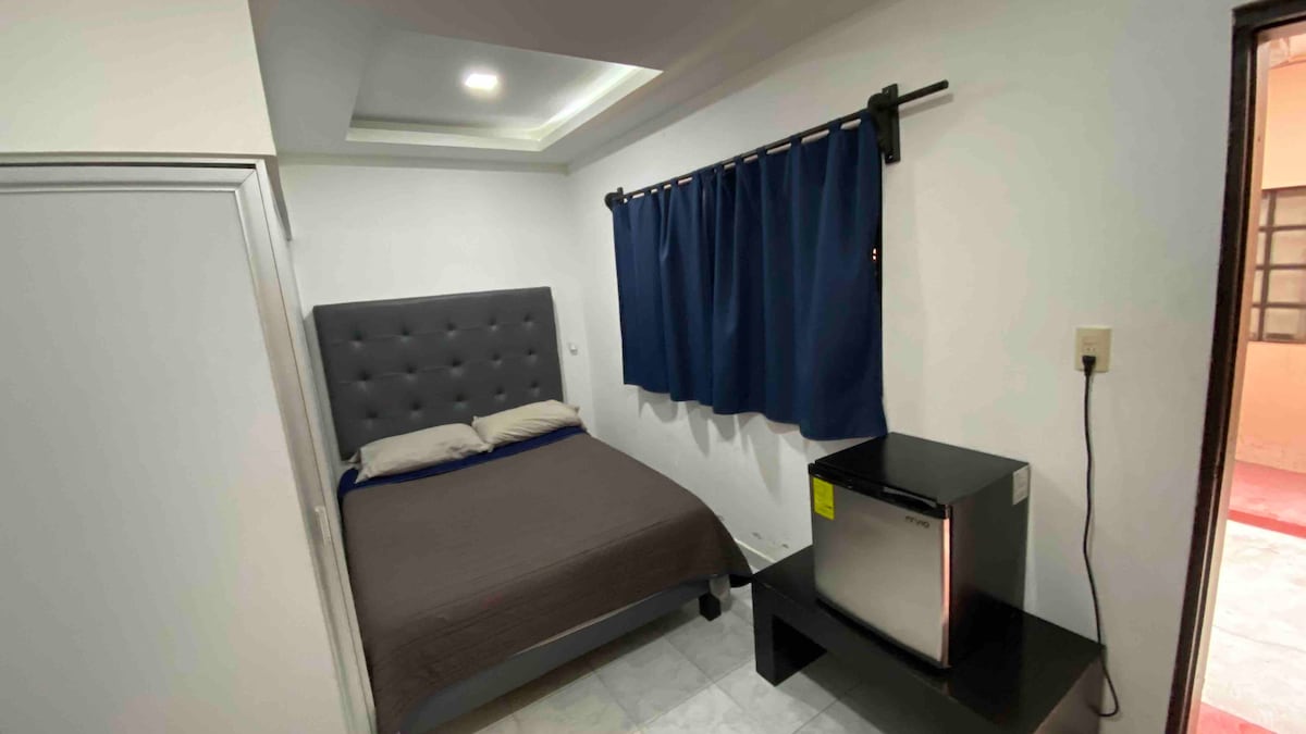 A simple bedroom features a comfortable bed with a gray upholstered headboard. Dark curtains frame a window, and a small black table holds a mini fridge. A light fixture is installed in the ceiling, providing illumination throughout the space.