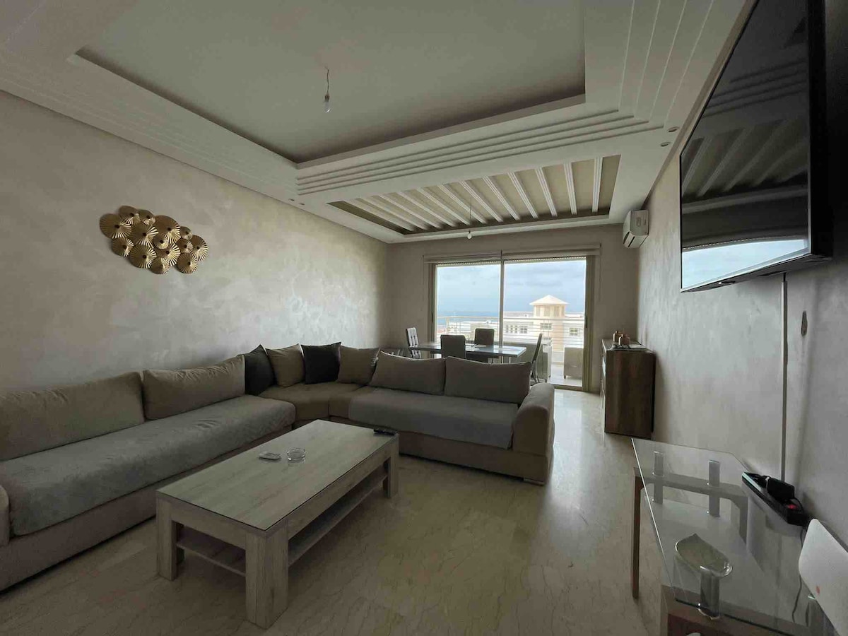 A spacious living area features a large sectional sofa, a glass coffee table, and a flat-screen TV mounted on the wall. Natural light fills the room through large windows leading to a balcony, offering a view of the surroundings.