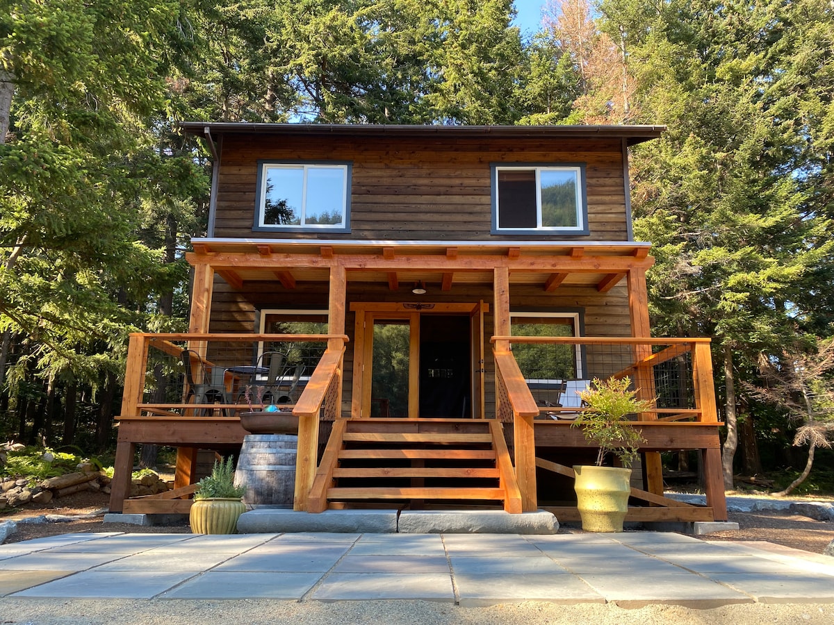 The exterior of a two-story wooden house is depicted, featuring a covered porch supported by sturdy wooden beams. Wide steps lead to the entrance, and large windows offer views of the surrounding trees. Potted plants are placed on the porch, enhancing the natural setting.