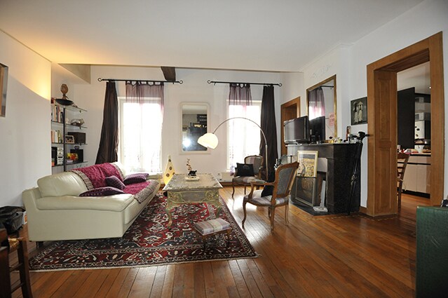 A spacious living area is showcased with a large, light-colored sofa and a patterned area rug. Two tall windows allow natural light to fill the room, which is adorned with a floor lamp and decorative shelving. A dark fireplace adds contrast to the bright décor.