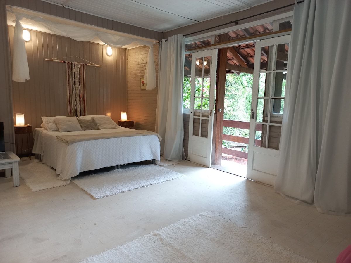 A spacious bedroom features a large, comfortable bed dressed in neutral tones. Soft rugs cover the floor, and large glass doors open to a tranquil outdoor space. Natural light fills the room, complemented by soft lighting from bedside lamps.
