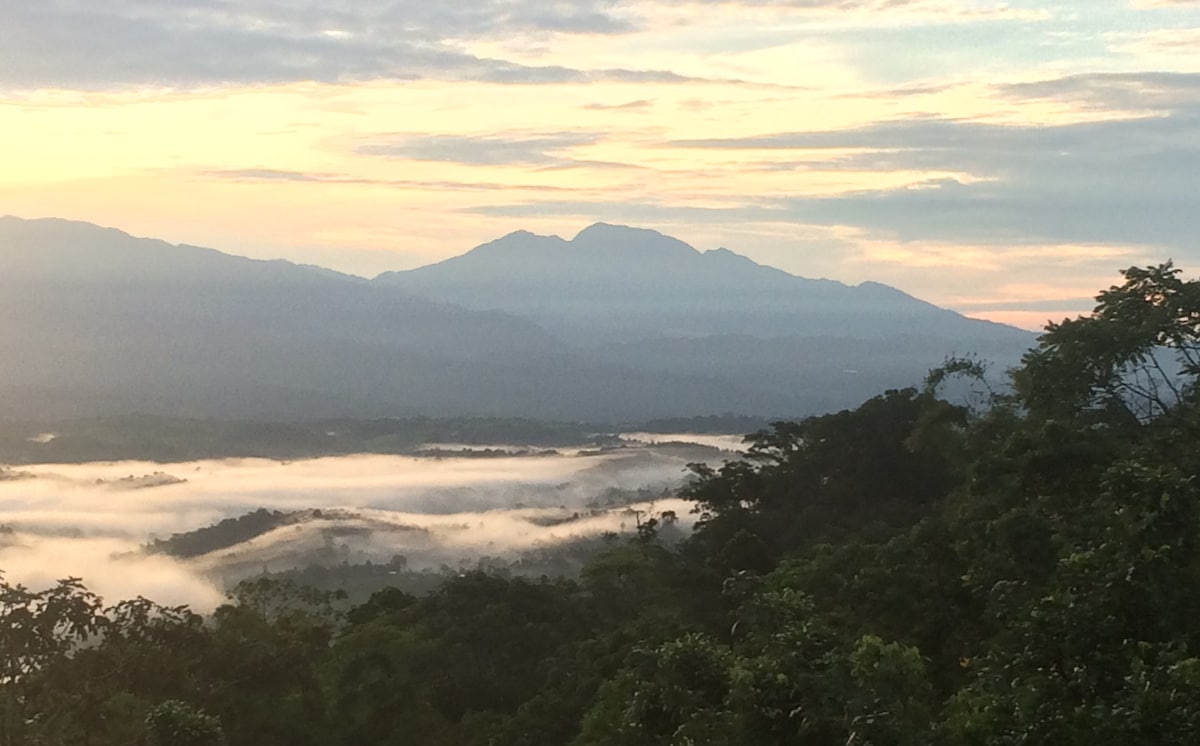 The image captures a panoramic view of the Talamanca mountain range at dawn. Soft clouds hover over lush valleys, creating a serene landscape illuminated by the gentle morning light. The vibrant greens of the surrounding trees frame the scene, enhancing the natural beauty.