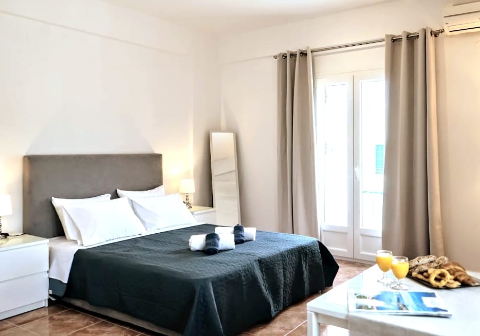 A minimalist sleeping area features a spacious double bed with quality bedding. Soft, neutral curtains frame the windows that lead to the balcony. A full-length mirror and two bedside lamps complement the decor, while a small table is set with drinks and reading materials.