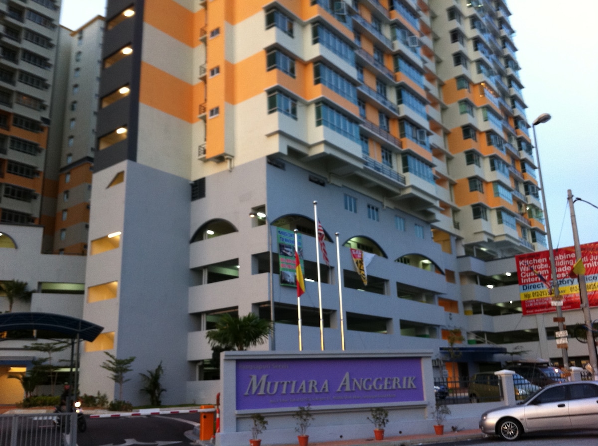 The exterior view of the Mutiara Anggerik apartment building is presented, featuring multiple floors with a combination of grey and orange facades. A large entrance sign is visible, along with flags gently waving in the breeze and landscaping that adds greenery to the environment.