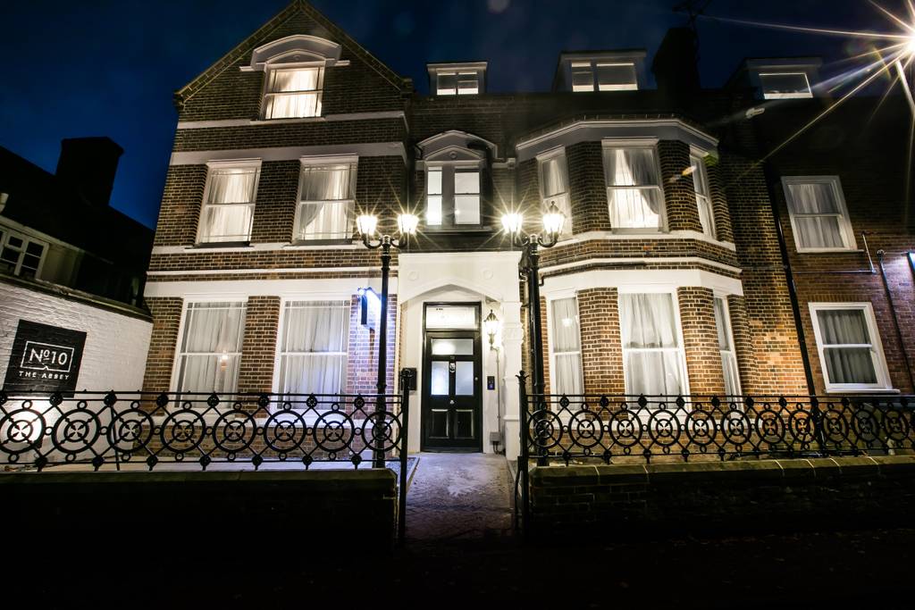 The exterior of No. 10 The Abbey features an elegant brick façade, complemented by large windows adorned with sheer curtains. The entrance is distinguished by a welcoming light fixture and a decorative wrought iron fence, illuminated softly by exterior lighting.