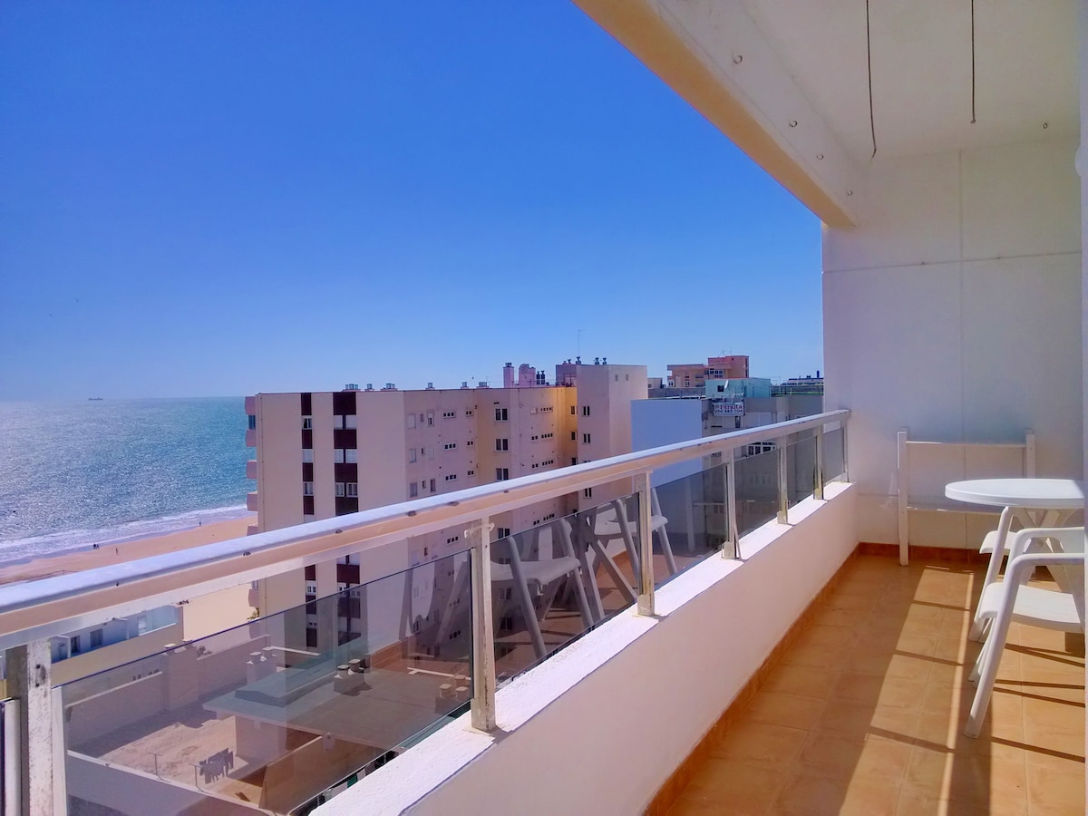 A sunlit terrace features a clear view of the beach and the ocean. The area includes a small round table and chairs, complementing the open space. The bright blue sky captures attention, while the neighboring buildings create a sense of the urban environment.