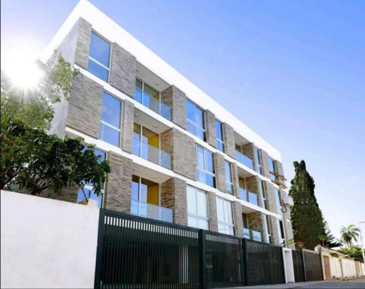 A modern four-story building features a combination of stone and glass exteriors. Large windows provide natural light, while balconies extend from each level. The entrance is framed by a sleek black gate, and lush greenery surrounds the property, enhancing the serene environment.