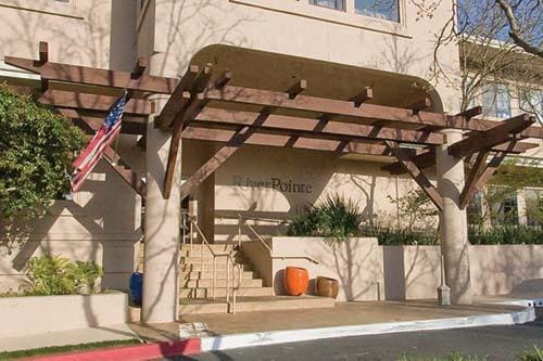 The entrance of Riverpointe Napa features a wooden pergola supported by concrete columns. The area is enhanced by a few potted plants and an American flag, while steps lead up to the lobby entrance. Natural light filters through the surrounding foliage.