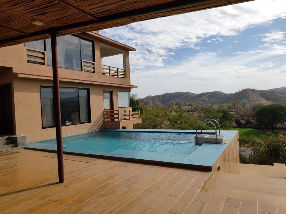 An outdoor area features an infinity swimming pool situated next to a spacious villa. The pool overlooks scenic rolling hills and lush greenery, with large glass windows reflecting the natural surroundings. Soft clouds drift across a bright sky.