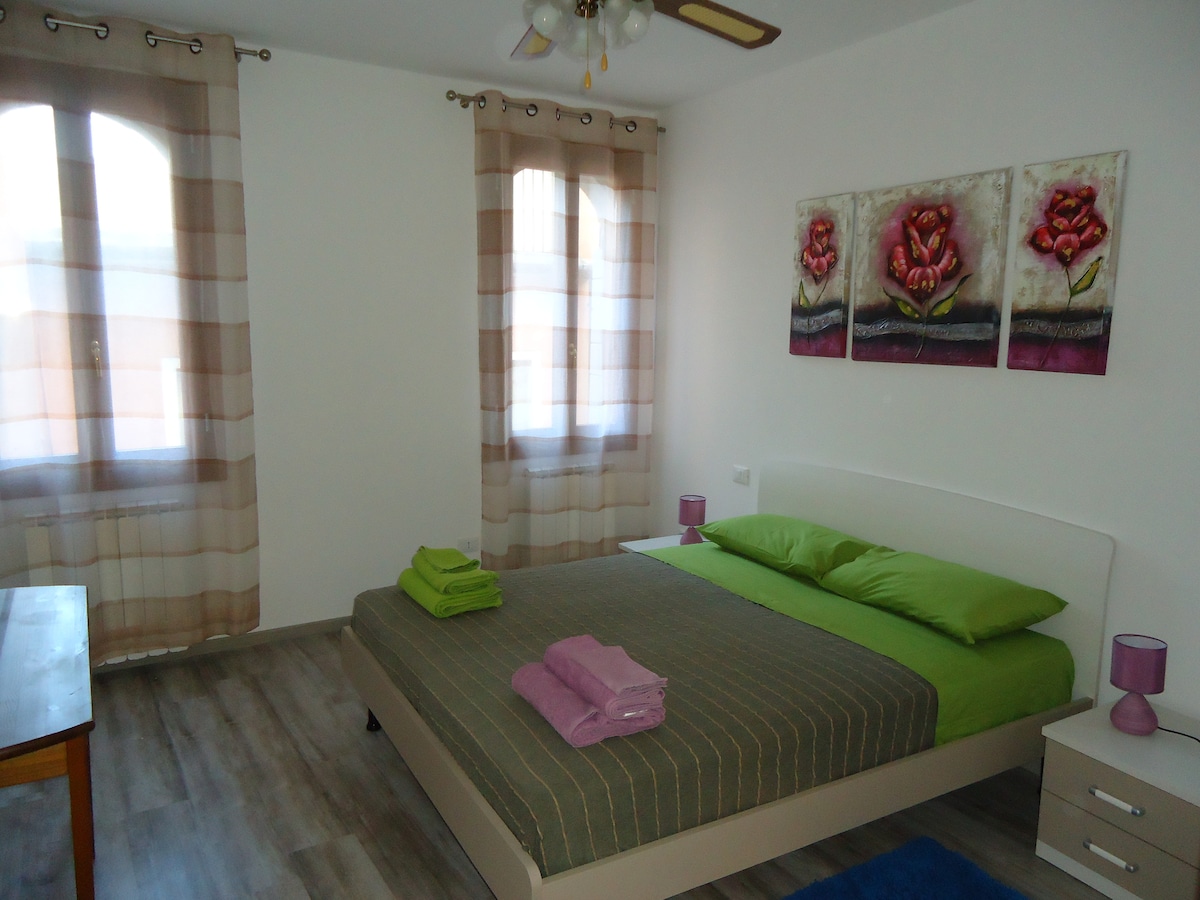 A spacious bedroom features a double bed dressed in green linens, accompanied by two decorative pillows. Nearby, a wooden table and a bedside table with lamps are visible. Large windows with sheer curtains allow soft light to illuminate the room, enhancing the colorful floral wall art.