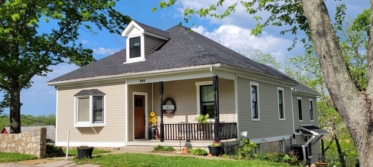 A charming single-story home features a gabled roof and a welcoming front porch adorned with sunflowers. Lush green grass surrounds the entrance, with trees providing shade. The property offers a blend of modern and historical elements, situated in a tranquil neighborhood.