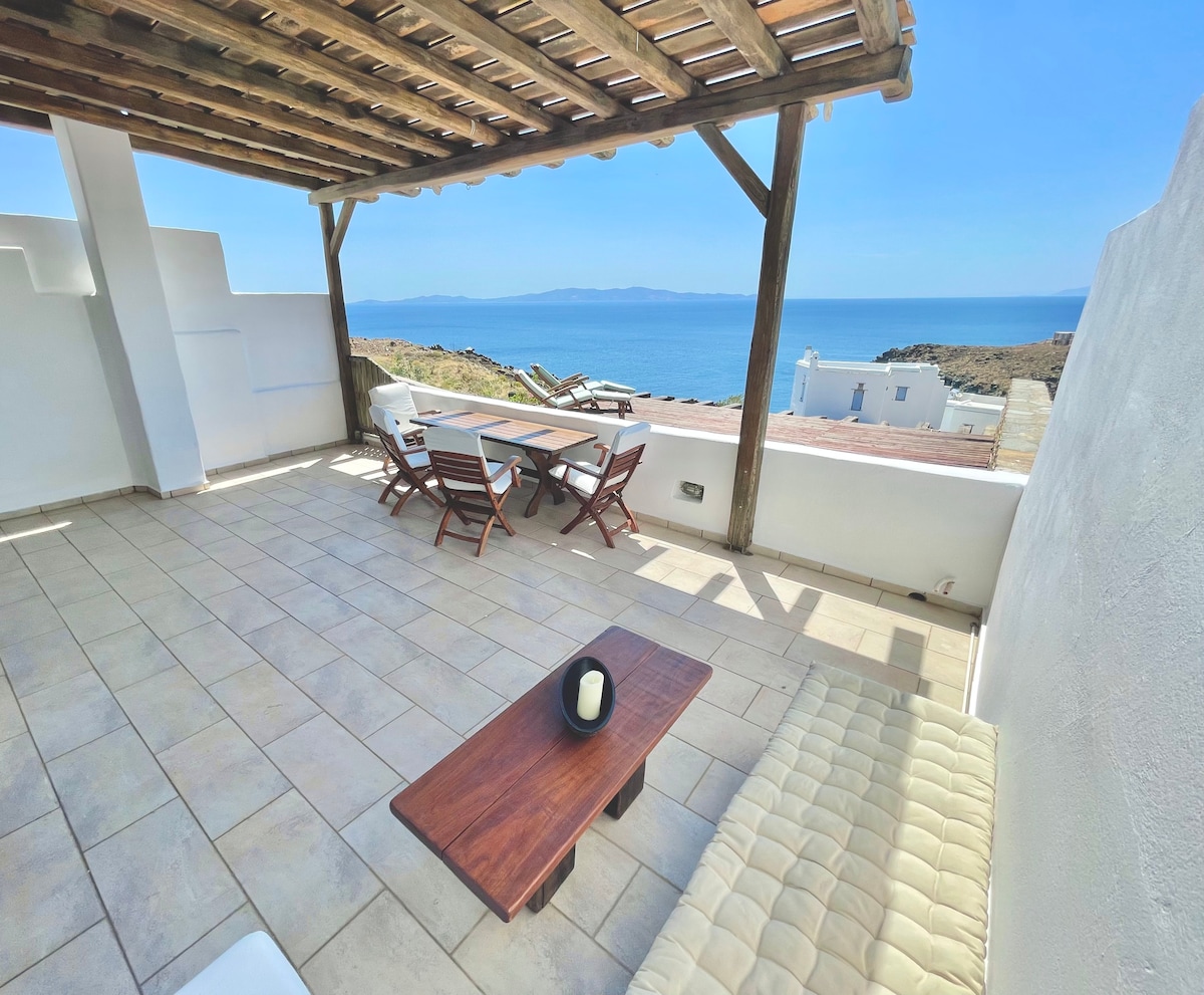 An expansive terrace features wooden beams overhead and a stunning sea view. A simple wooden coffee table is positioned near a comfortable seating area with four chairs. The floor is tiled, creating a clean, open space perfect for relaxation.