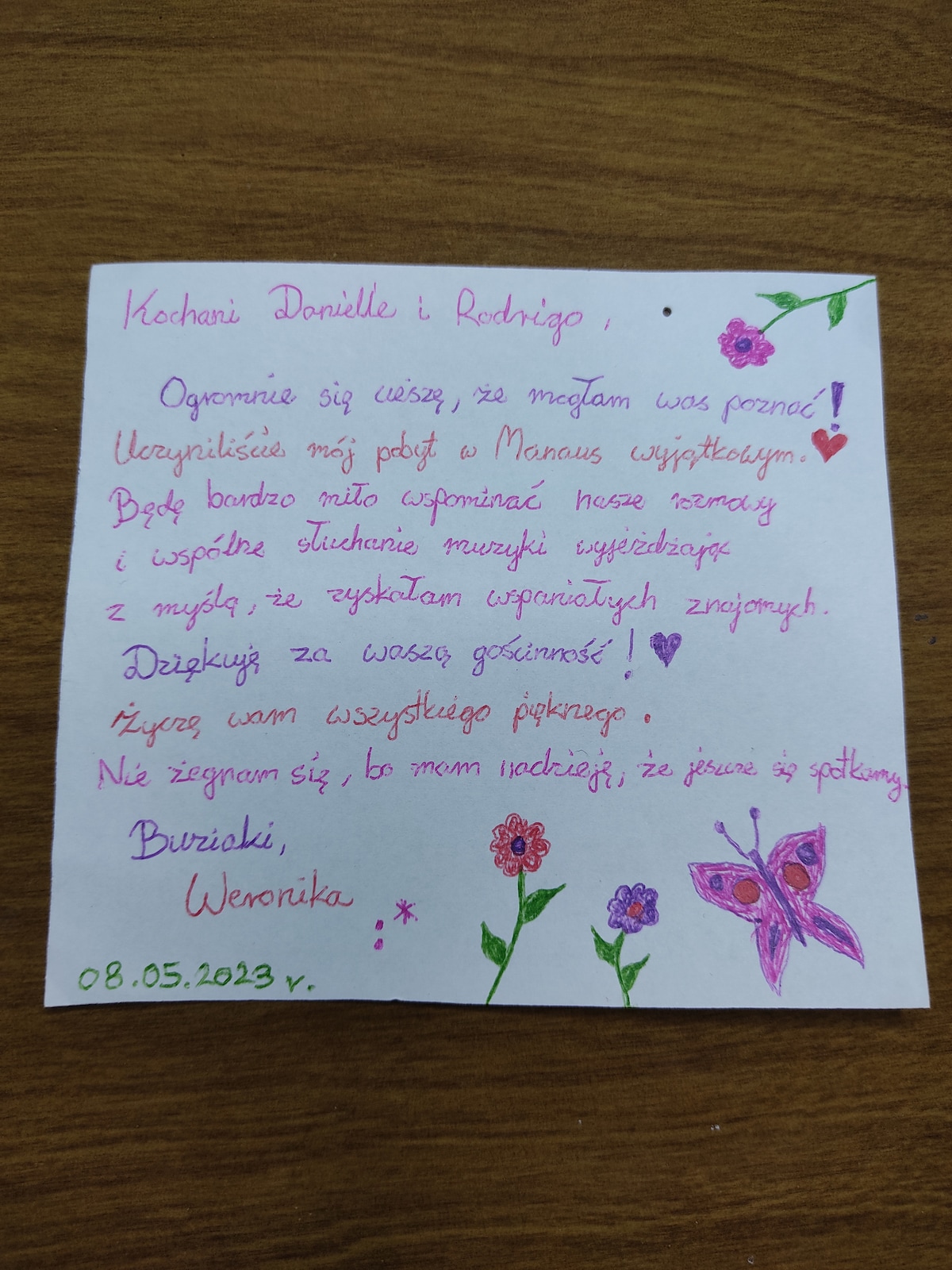 A handwritten note is displayed on a wooden surface. The note features colorful drawings of butterflies and flowers, enhancing its personal touch. The text is written in a cheerful, flowing script with a mix of pink and purple ink, conveying warm sentiments and gratitude.