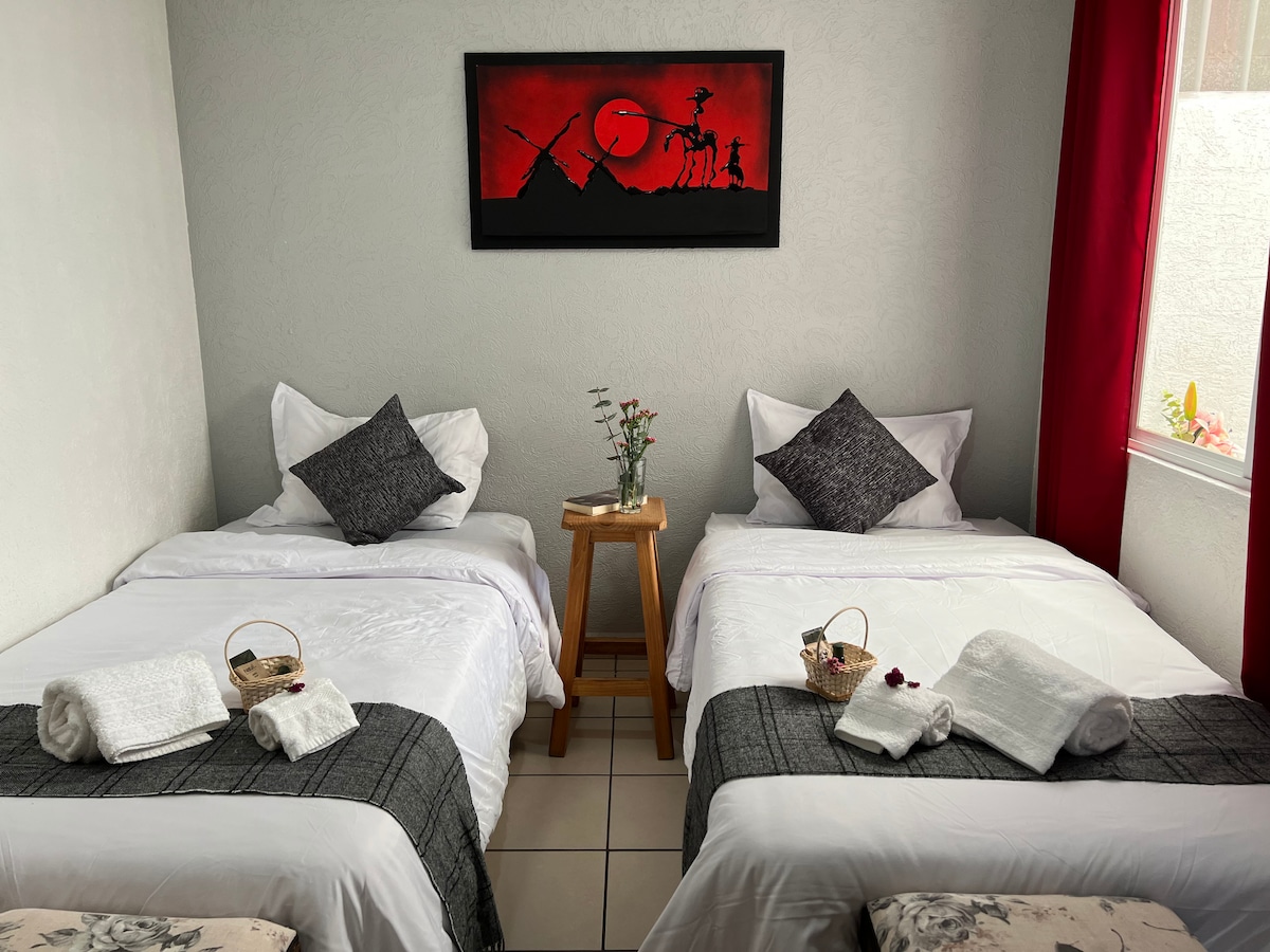 A cozy sleeping area features two neatly made beds with crisp white linens and gray throw blankets. Decorative pillows add a touch of style. A small wooden table holds a flower arrangement, while light enters through a window adorned with red curtains.