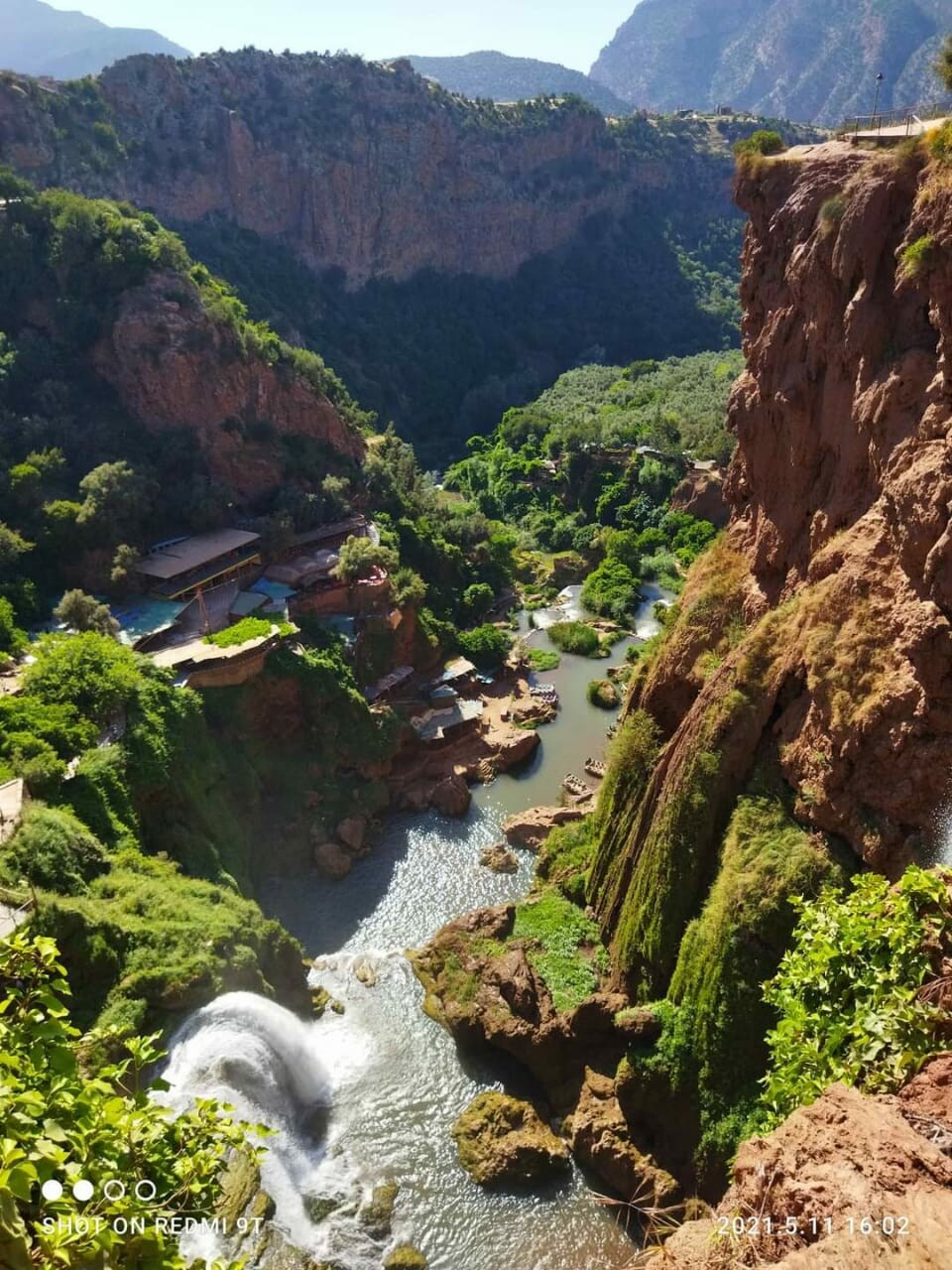 A scenic view captures a lush green landscape shaped by rocky cliffs and a flowing river. Waterfalls cascade down the rocky terrain, creating a serene atmosphere. Surrounding vegetation enhances the natural beauty of the area, while distant mountains frame the horizon.