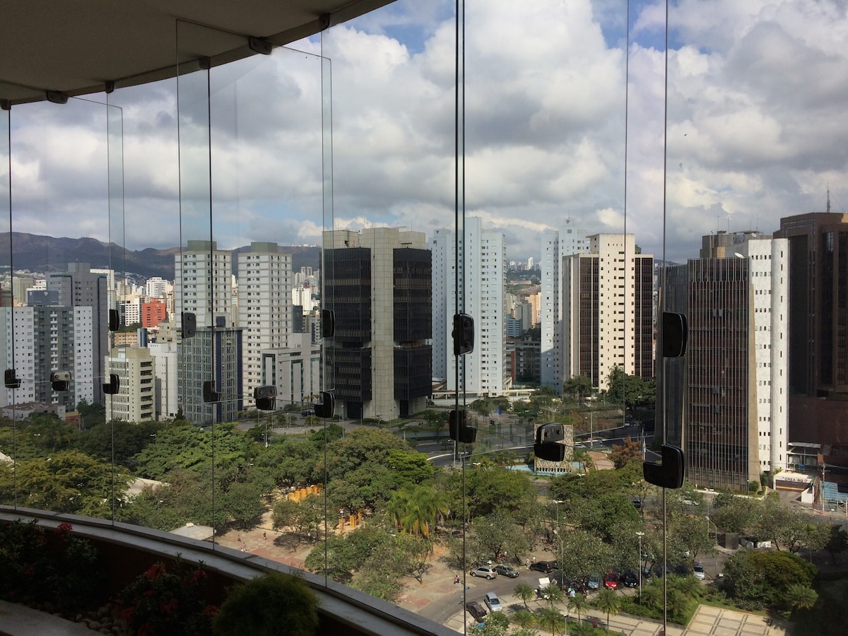 A panoramic view showcases a skyline filled with tall buildings and lush greenery below. The cityscape is framed by large glass windows, reflecting the sky with clouds, creating an expansive feeling of openness.
