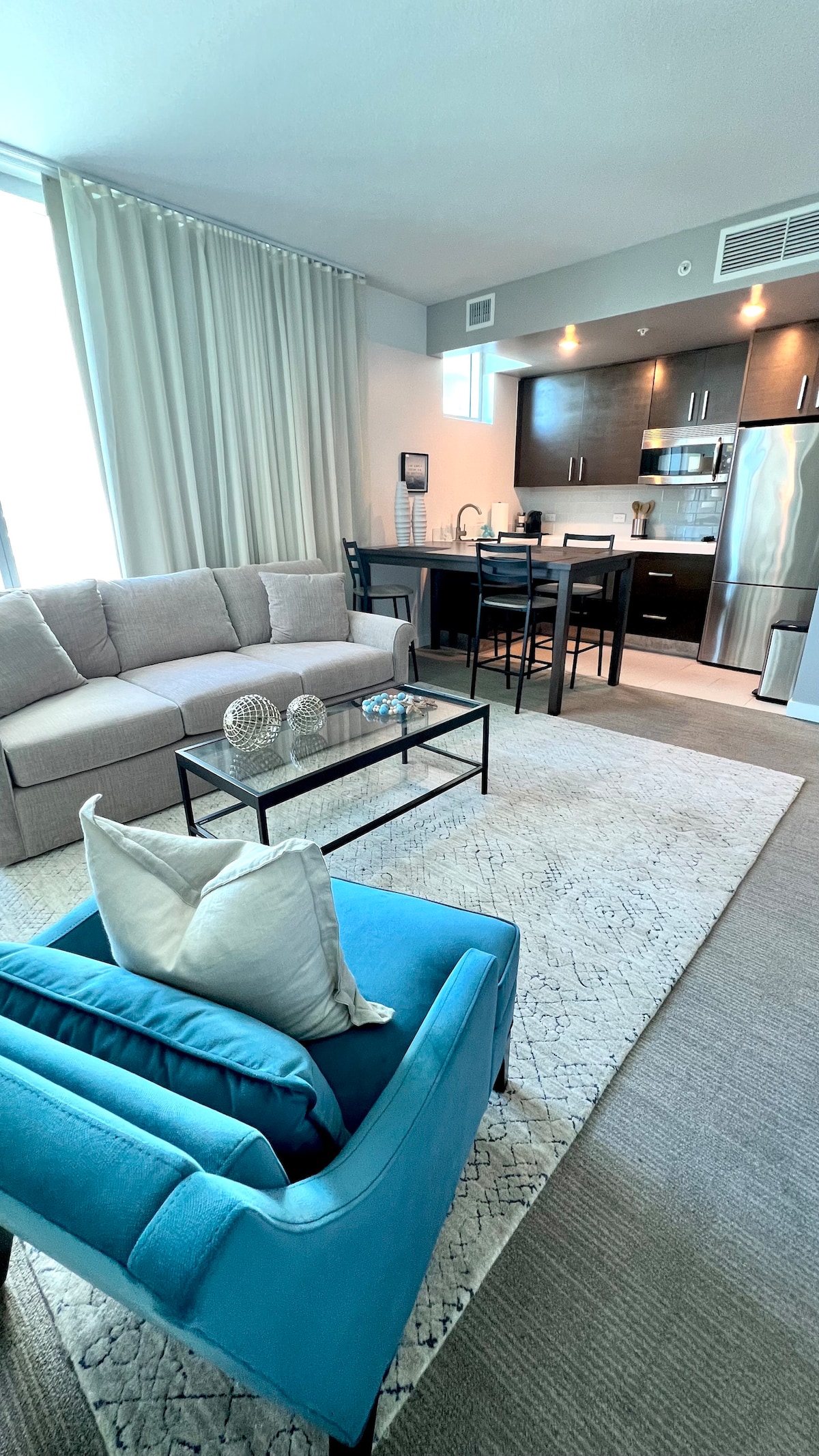 A light-filled living area features a spacious sectional sofa in neutral tones and a colorful blue armchair. A glass coffee table sits atop a soft area rug, with decorative items displayed. The modern kitchenette is visible in the background, showcasing dark cabinetry and stainless appliances.