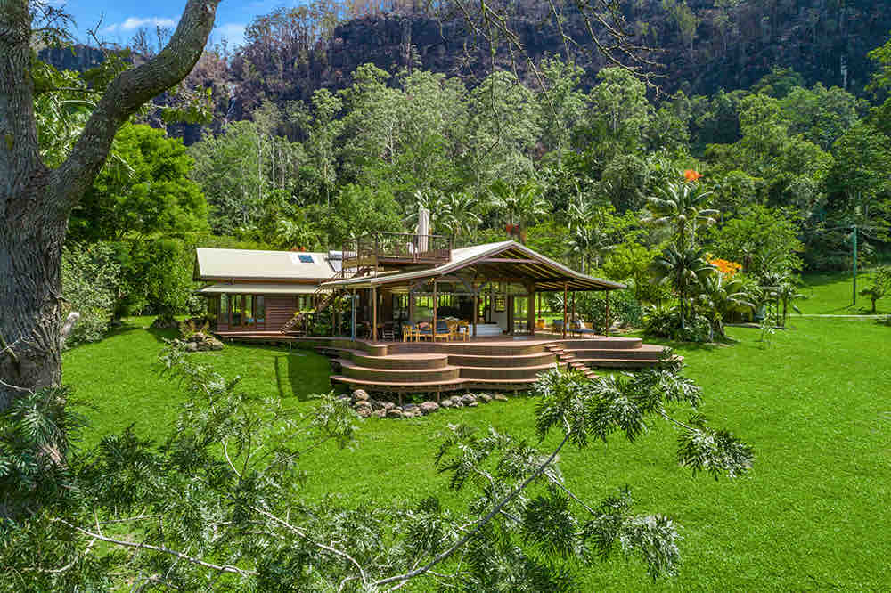 The house is nestled within a lush green landscape, surrounded by tall trees and rugged cliffs in the background. Large decks extend from the building, providing ample outdoor space and a connection to nature. The area is peaceful, showcasing the serene environment.