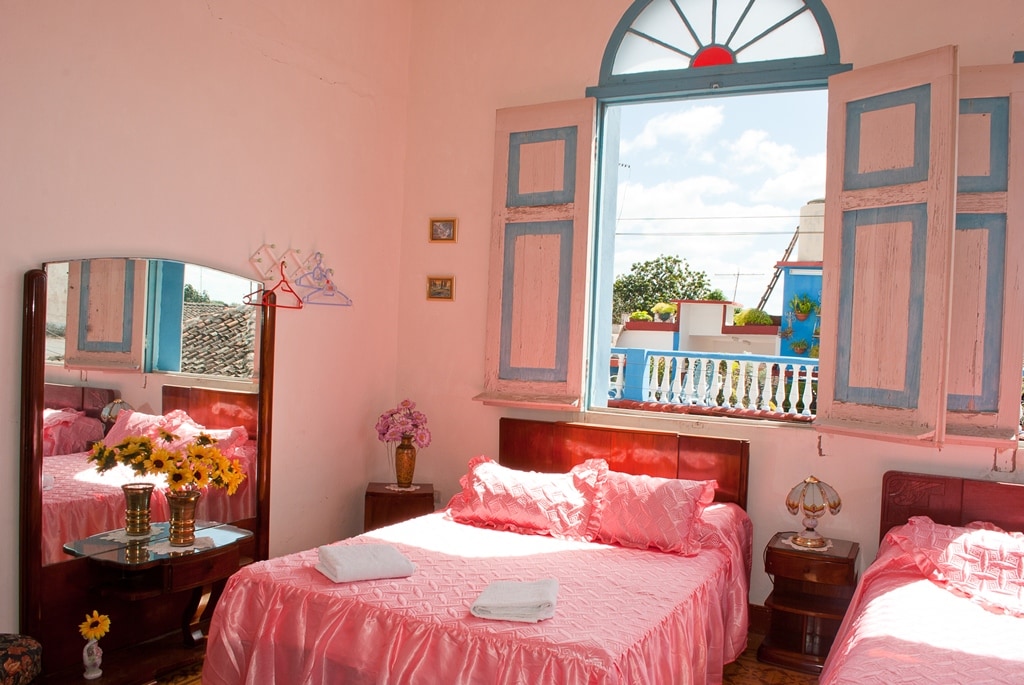 A guest room features two spacious beds dressed in soft pink linens, complemented by a decorative mirror. Natural light enters through an arched window, framed in blue, overlooking a glimpse of the colorful exterior. Fresh flowers in a vase add a touch of charm.