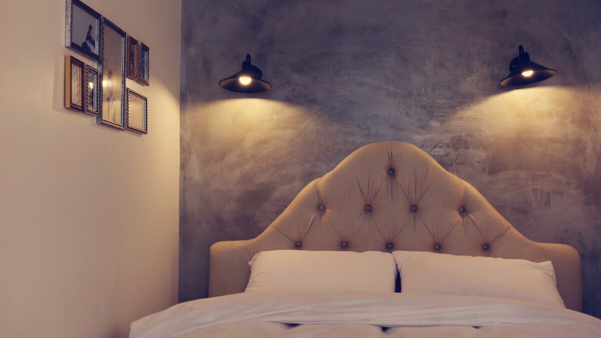 A stylish sleeping area features a tufted headboard with soft lighting on either side. Neutral-toned walls are complemented by framed wall art, contributing to a modern yet cozy ambiance. Crisp white bedding is neatly arranged on the spacious bed.