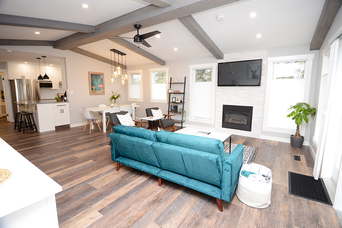 A spacious living area is designed with a teal sofa and wooden beam ceilings, allowing for a cozy yet modern atmosphere. A natural gas fireplace is present, accompanied by a wall-mounted television. The open layout connects to a bright dining area, enhancing the communal space.