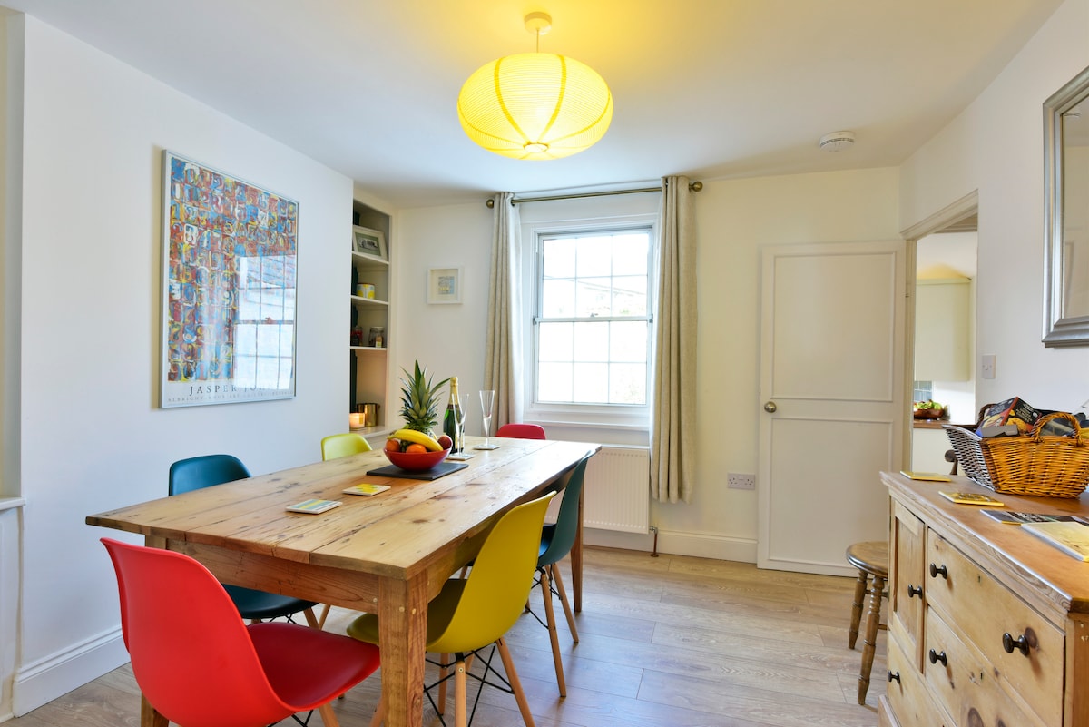 The dining area features a reclaimed Victorian pine table surrounded by colorful Eames-style chairs. Natural light enters through a large window, while white walls and light wood flooring create a bright and airy atmosphere. A modern light fixture hangs above the table.