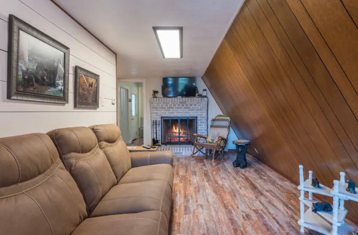 A living area features a warm-toned leather sofa positioned beside a traditional fireplace with a brick surround. A mounted television is visible above the fireplace, while artwork hangs on the wall. The sloped ceiling adds architectural interest to the cozy space.