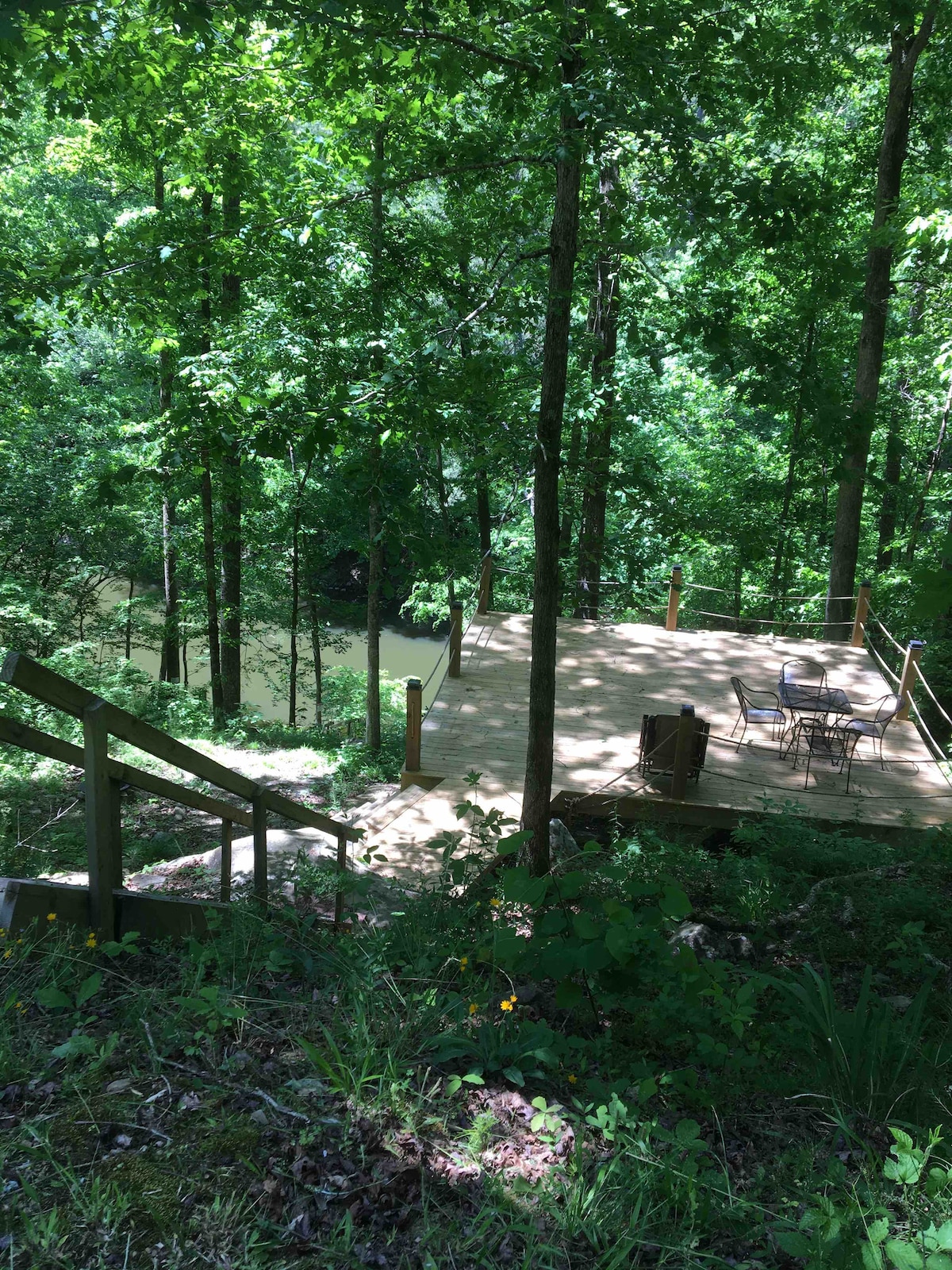 A wooden deck is nestled among tall trees, overlooking a serene body of water. Outdoor seating, including a table and chairs, is positioned on the deck, inviting relaxation amidst the lush greenery. A set of stairs leads down from the cabin to the deck area.