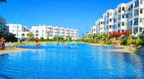 A large swimming pool is surrounded by contemporary apartment buildings with white facades. Vibrant greenery and colorful flowers frame the pool area, where guests can be seen enjoying the water in a relaxed atmosphere. Clear blue skies enhance the tranquil setting.