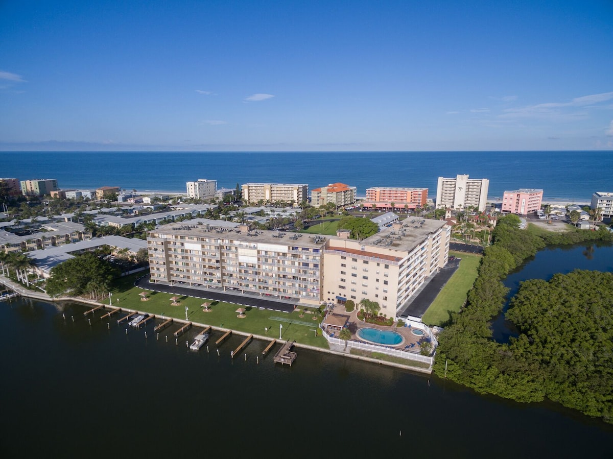 An aerial view of the Bay Shores Yacht and Tennis Club showcases the building’s location by the waterfront. The surrounding landscape includes green areas and nearby accommodations, with the expansive beach and ocean visible in the distance under a clear blue sky.