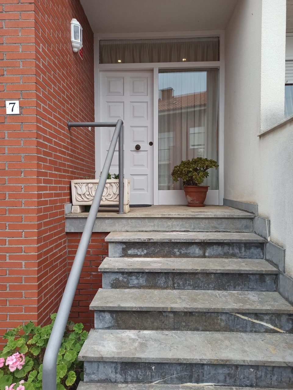 A set of stone steps leads to a white front door, framed by a curtain behind glass panels. A potted plant sits beside the door, and a decorative stone piece is positioned on the ground, adding a welcoming touch to the entryway.
