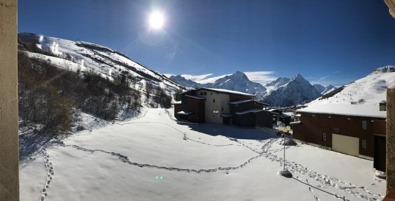A panoramic view captures a snow-covered landscape featuring diverse mountain ranges under a clear blue sky. A residential building is visible in the foreground, surrounded by untouched snow. Footprints lead into the snowy area, suggesting access to outdoor activities.