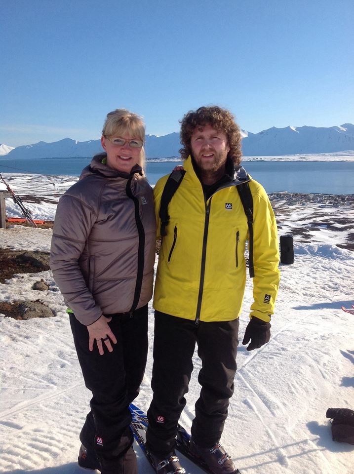 Two individuals are standing on a snowy landscape, wearing jackets suited for cold weather. The backdrop features a scenic view of distant mountains and a body of water under a clear blue sky.