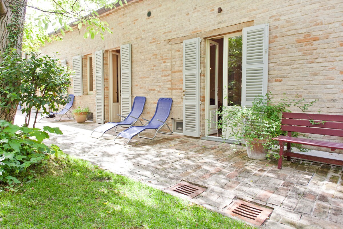 A serene outdoor area is featured, with lounge chairs positioned on a brick pathway. Green foliage surrounds the space, while potted plants add natural elements. Light-colored walls and open shutters create a welcoming atmosphere, inviting guests to relax in the garden.