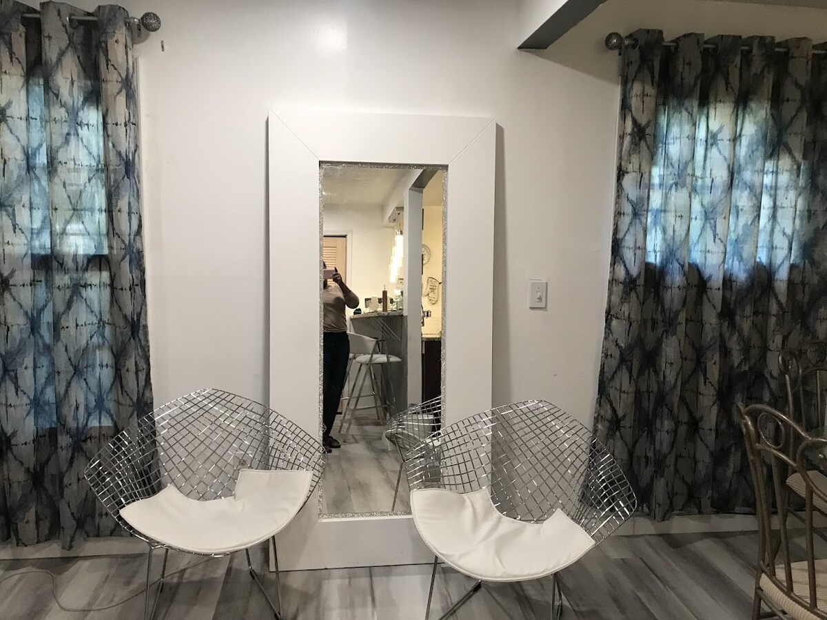 Two wire chairs with white cushions are positioned in front of a large, rectangular mirror. The mirror reflects a bright area, while patterned curtains frame the space, adding privacy and style. Light filters through the window, enhancing the inviting ambiance.