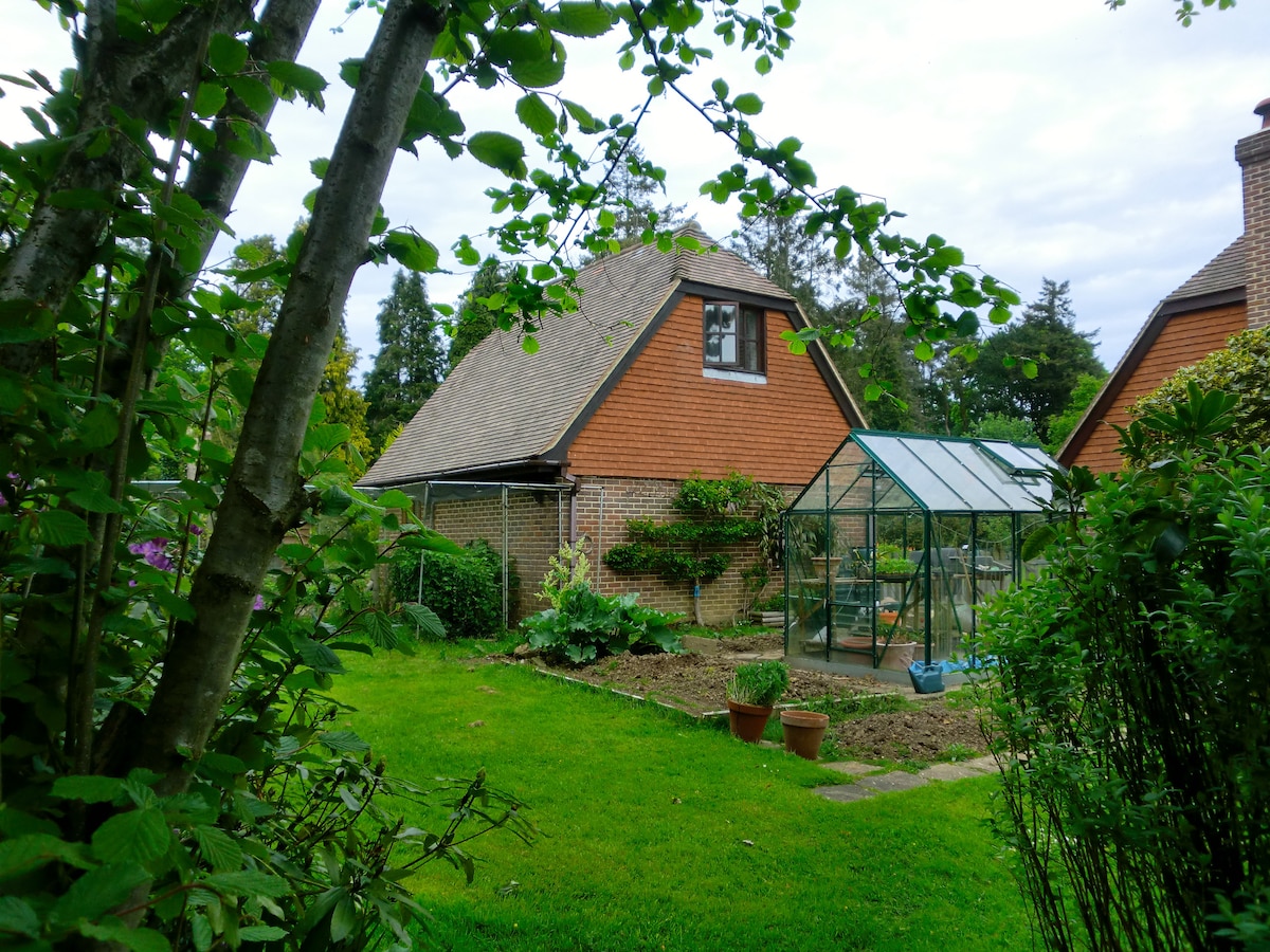 The exterior of a detached loft accommodation is shown, framed by lush greenery. A greenhouse is visible nearby, surrounded by a well-maintained garden, featuring pots and a spacious lawn. A charming thatched roof complements the natural setting, evoking a serene outdoor atmosphere.