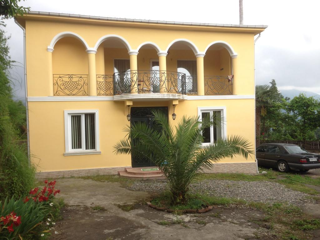 A two-story yellow building features arched windows and a decorative balcony. Lush greenery surrounds the entrance, with a well-defined pathway leading to the front door. A parked car is visible on the gravel driveway, emphasizing the access to the property.