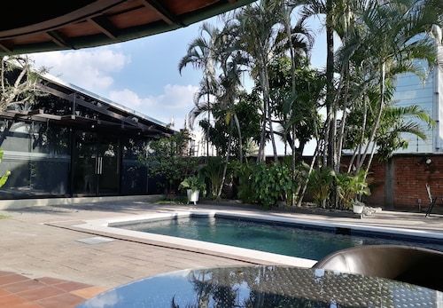 A serene outdoor area features a swimming pool surrounded by lush greenery and tropical plants. The space includes modern architecture with large glass doors, providing a seamless connection between indoor and outdoor environments.