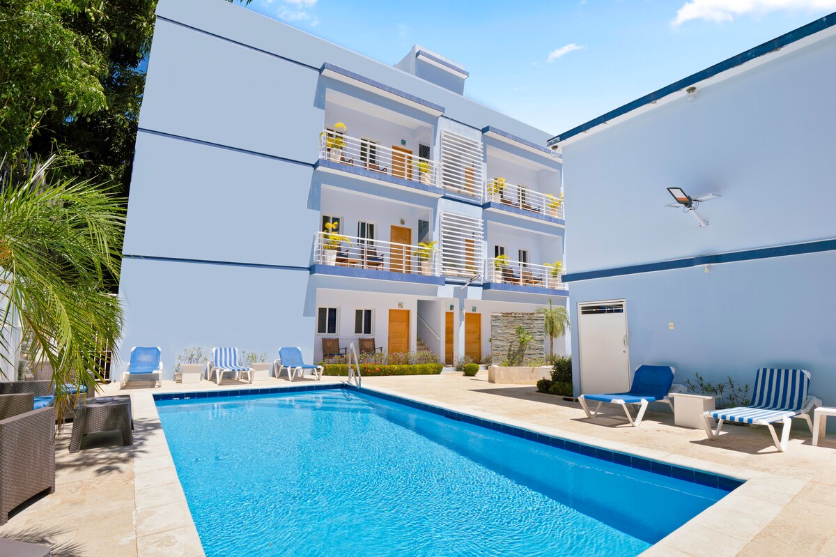 The image showcases a modern building surrounding a refreshing pool area. Sun loungers are arranged near the pool, providing comfortable seating. Balconies with bright yellow chairs are visible on the upper levels, enhancing the inviting atmosphere of the complex.