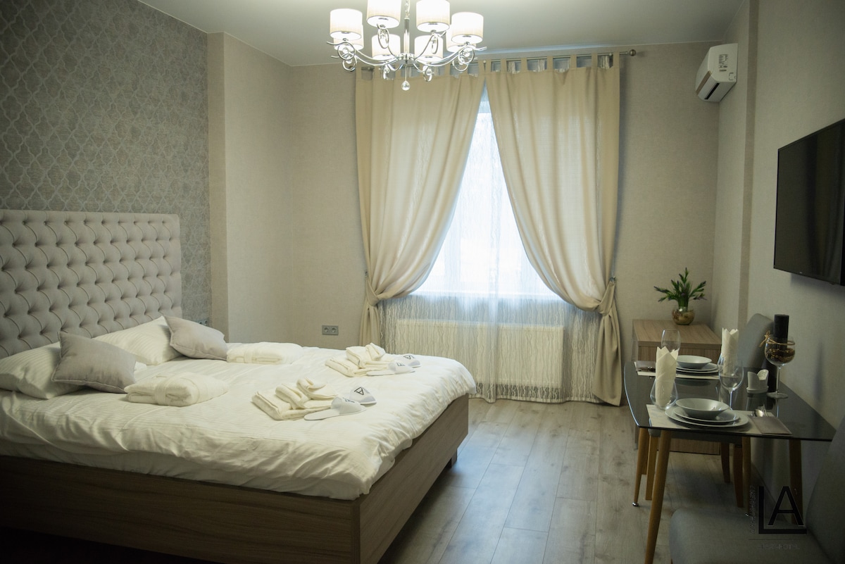 A spacious bedroom features a large bed with neatly arranged linens and several towels. Natural light enters through a window covered with sheer curtains. A small dining table is set up for two, complemented by an elegant chandelier above, contributing to a cozy environment.
