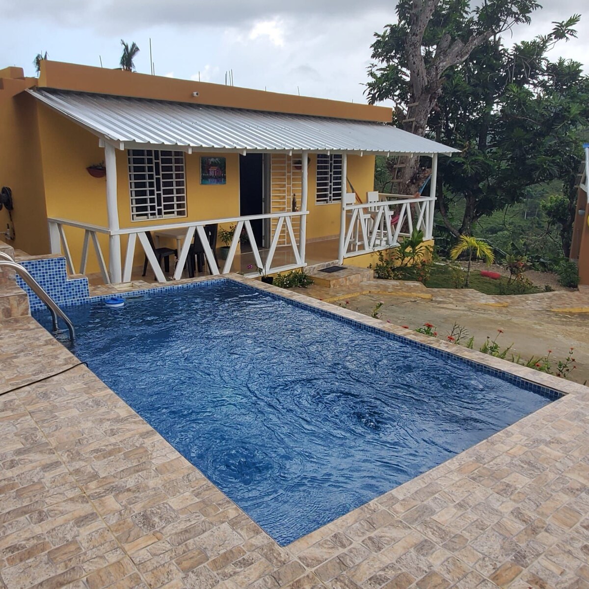 A private swimming pool is surrounded by a tiled terrace, complemented by lush greenery. The house features a covered porch with white railings, providing a charming view of the landscape and inviting relaxation.