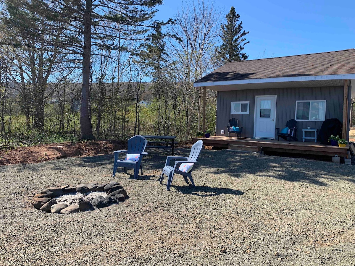 A cozy cabin is situated near a gravel area featuring a circular fire pit surrounded by stone. Two blue chairs are placed invitingly in front of the cabin, which is set against a backdrop of trees and open green space.