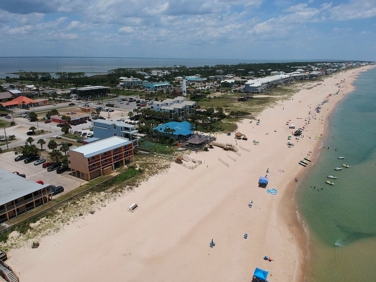 The beachfront area of St. George Island is showcased, featuring a wide stretch of sandy shore lined with colorful umbrellas and beachgoers. Buildings and palm trees can be seen in the background, with the blue Gulf of Mexico gently lapping at the shoreline.