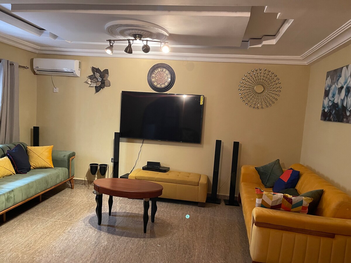 A spacious sitting room is equipped with a large smart TV, two sofas in different styles, and a round coffee table. Wall decor includes artwork and mirrors, while natural light filters in through the curtains. Air conditioning units ensure comfort within the space.