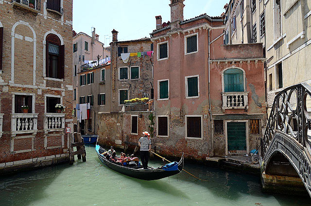 A traditional gondola glides through a narrow canal, flanked by historic buildings. The facades feature a mix of textured brick and painted surfaces, with green window shutters adding color. Laundry hangs between buildings, while a charming bridge offers a glimpse of the street beyond.