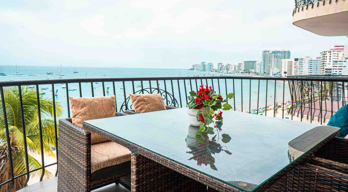 A spacious balcony is visible, equipped with a woven table and comfortable seating. A potted plant with vibrant red flowers adds a touch of color. Expansive views of the ocean and surrounding buildings are framed by the balcony railing.