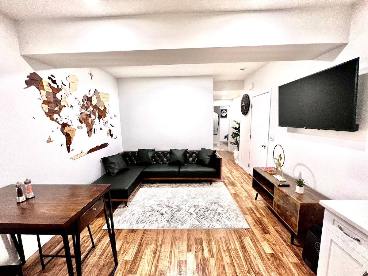 A modern living area features a dark sectional sofa positioned along a wall adorned with a wooden world map. A rectangular wooden table sits in the foreground, complemented by a patterned area rug beneath. A television is mounted on the opposite wall.