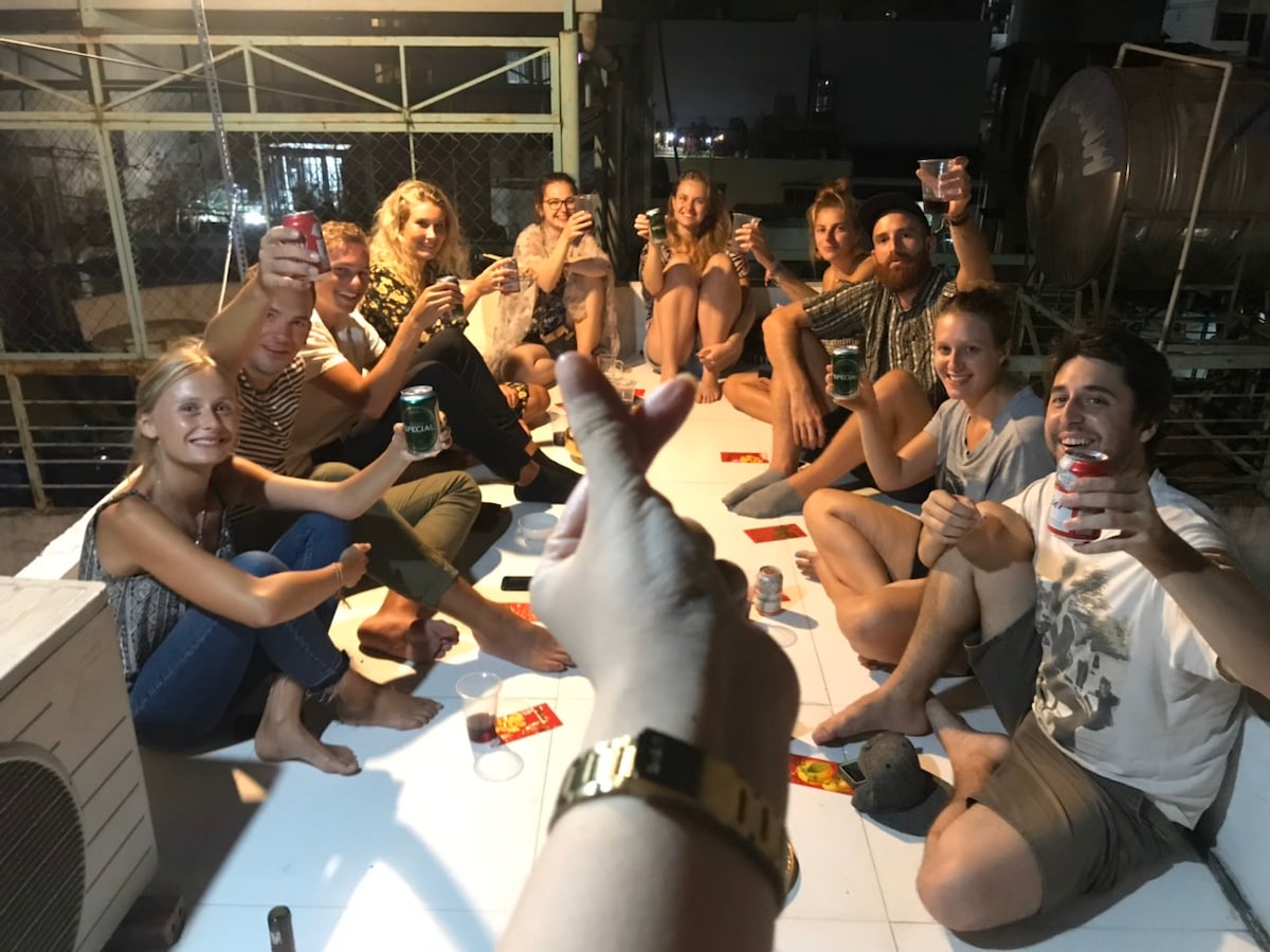 A lively gathering is captured on a rooftop, featuring a group of individuals seated in a circle. Drinks are raised in celebration, with an array of beverages visible. The scene is warmly lit, suggesting a relaxed atmosphere. The surrounding structure includes metal supports and outdoor seating.
